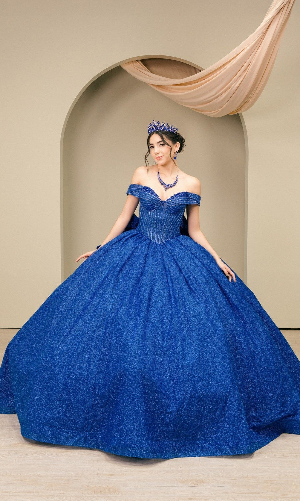 A woman wearing the Quinceanera Dress 8460JQ By Cinderella Couture in royal, with a matching necklace and tiara, stands before a beige arched backdrop draped with fabric.